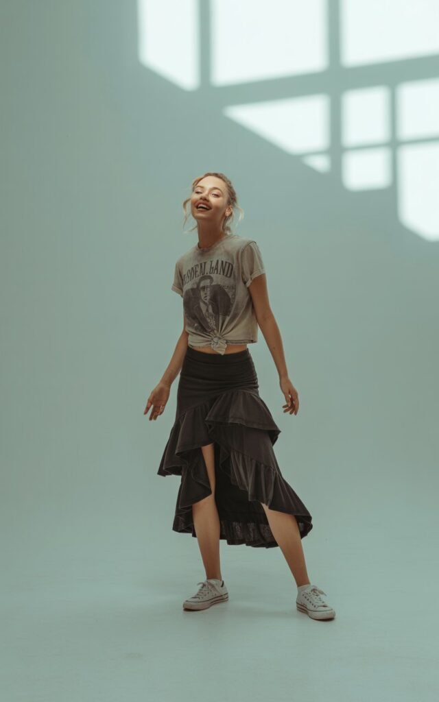 Full-body fashion shot in a minimalist studio. Model wears a vintage band tee tucked into a layered ruffled black skirt and sneakers. Soft gray background and natural light. Pose playful with slight movement in skirt. Expression fun, leaning slightly forward with energy.