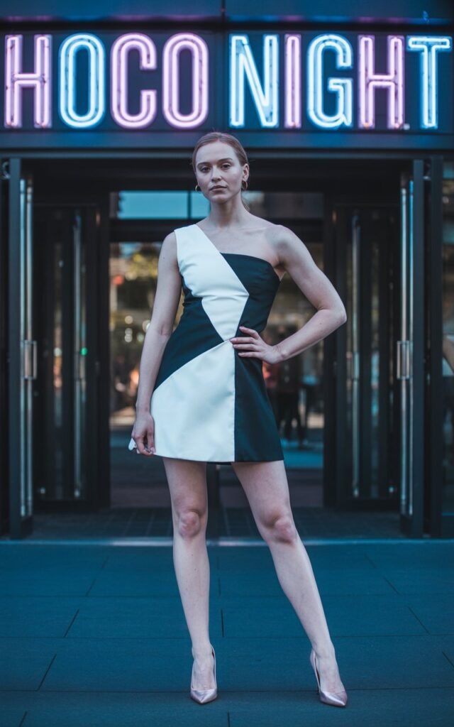 Full-body image of a white-skinned girl in a high-contrast two-tone mini dress, structured silhouette, and matching heels. Photographed in front of the gym entrance with neon signage for hoco night. Cool-toned lighting highlights the color contrast. Confident pose, natural skin details.
