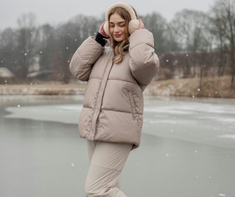Full-body image of a white-skinned girl in a matching beige puffer jacket and pants, plush ear muffs, and winter boots. She’s standing on a frozen pond edge with light snowfall. Soft overcast light emphasizes monochrome tones. Pose smiling while holding earmuffs, casual relaxed stance.