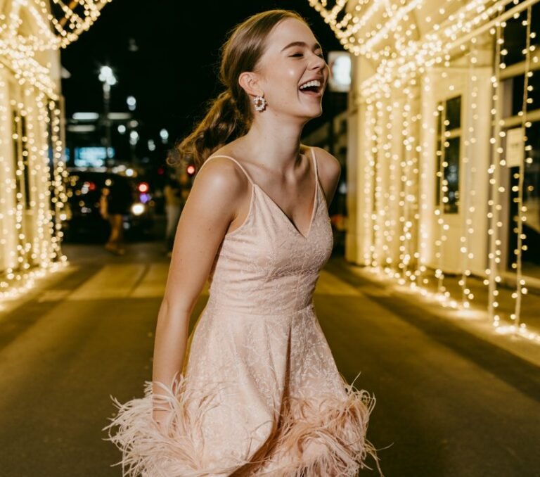 Full-body image of a white-skinned girl wearing a feather-trim mini dress in soft white or blush, playful earrings, and platform heels. She’s captured mid-step near the hoco entrance arch covered in fairy lights. Slight movement in feathers adds texture. Soft event lighting and candid laughter.