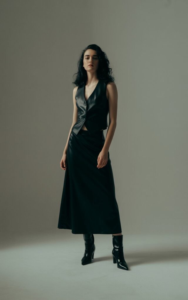 Full-body image of a white-skinned model with long silky dark hair, wearing a black tailored vest and matching black midi skirt with heeled boots. Dramatic indoor studio lighting with soft shadows. She stands tall with a poised, elegant expression. Clean, powerful look.