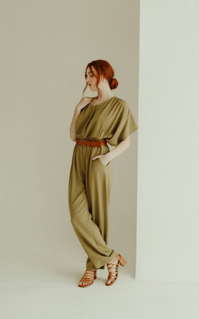 Full-body image of a white-skinned woman with auburn hair in a low bun, standing on a minimalist studio set with soft daylight. She wears an olive-green relaxed jumpsuit cinched with a brown belt, and tan sandals. One hand rests in her pocket, the other adjusting her hair. Clean, modern background enhances the outfit’s ease.