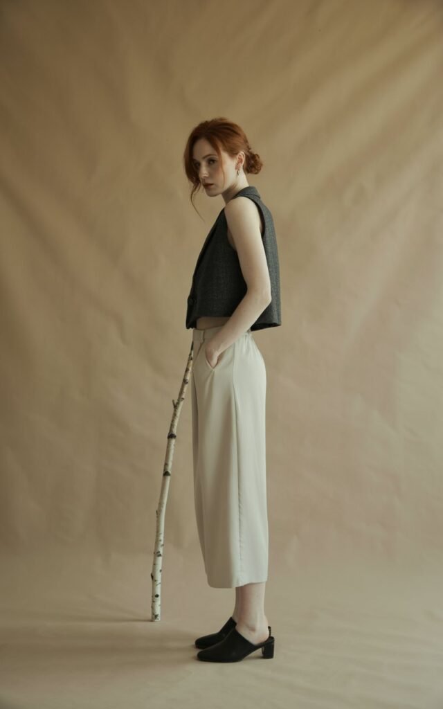 Full-body image of a white-skinned woman with auburn hair in a messy bun, wearing a charcoal cropped vest and cream high-waist culottes, styled with black mules. Studio setting with minimalist beige backdrop and soft diffused lighting. She stands in a slight contrapposto pose. Calm, confident expression.