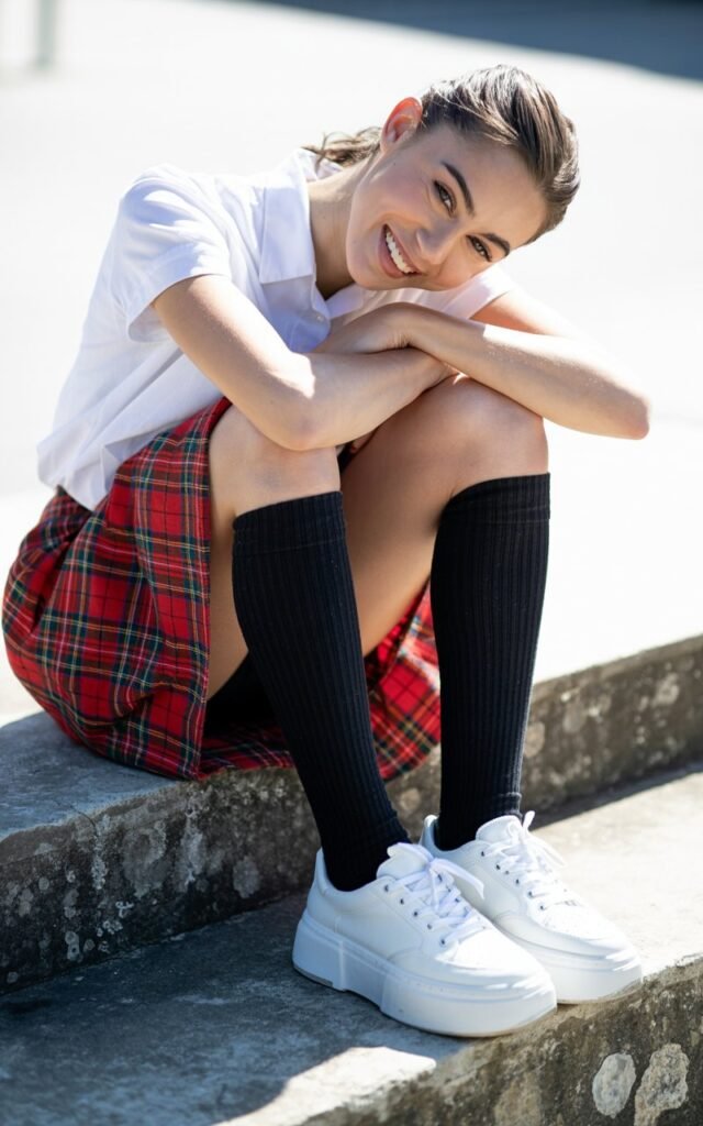Full-body outdoor photo on concrete steps. Model wears a plaid skirt, black knee-high socks, and white platform sneakers. Natural daylight with bright contrast. Sitting casually, leaning on knees with a playful smile. Youthful energy, clean sharp details in texture and fabric.