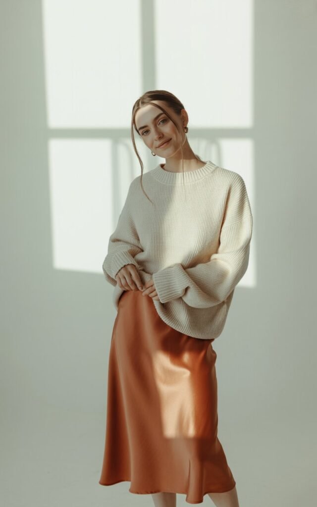 Full-body photo of a white-skinned model with relaxed hair, wearing a cream chunky knit sweater and a rust-colored satin skirt. Clean indoor studio with soft window light. She stands with a gentle smile and relaxed posture. Clear contrast between cozy knits and glossy satin for realism.