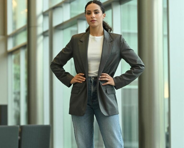 Full-body shot in a modern office lobby. She wears a gray tailored blazer, white tee, medium-wash straight jeans, and pointed flats. Hair is in a smooth ponytail. Clean indoor lighting with natural highlights. She stands with hands on hips, confident and polished.