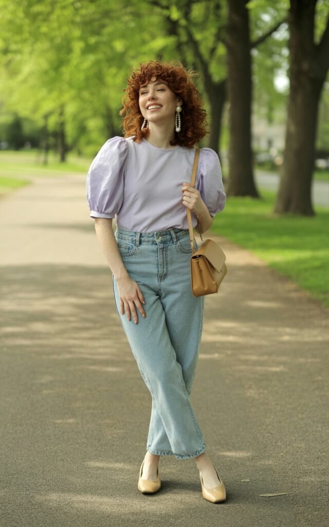 Full-body shot of a white-skinned female model with curly shoulder-length hair, standing in a park. She wears a pastel puff-sleeve blouse tucked into light-wash high-rise jeans, with flats and pearl earrings. Natural daylight. Her expression is warm and relaxed, gently holding her bag at her side.