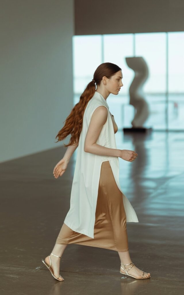 Full-body shot of a white-skinned woman with long chestnut hair in a low ponytail, wearing a white longline vest over a tan column dress with strappy sandals. Captured in a modern art gallery with bright diffused natural light. She walks gracefully, dress flowing slightly. Elegant, serene expression.
