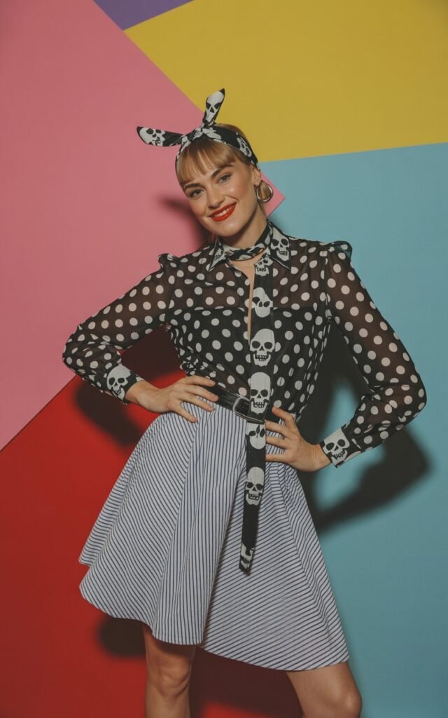 Full-body studio photo with patterned backdrop. Model wears a polka-dot blouse, striped skirt, and skull-printed accessories. Mix of bold textures and fabrics. Soft, indirect lighting. Playful yet edgy pose — hands on hips, smirk with attitude. Vibrant pop-art aesthetic.