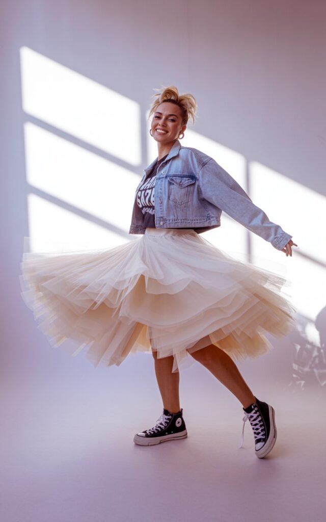 Full-body studio portrait of a model in a layered white tulle skirt, graphic tee, cropped denim jacket, and chunky black Converse. Hair half-up, teased volume. Soft window lighting from the side. She’s mid-twirl, skirt flowing. Expression playful yet confident, surrounded by subtle pastel tones.