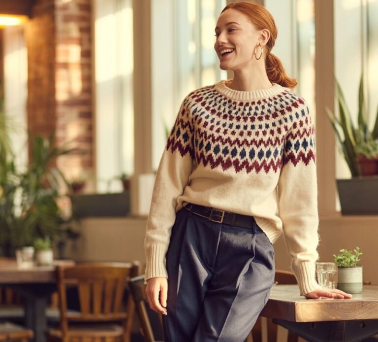 Full-body view of a model in a patterned fair isle sweater with solid navy trousers, black loafers, and small hoop earrings. Cozy café interior with warm natural light. Hair in a low ponytail, subtle makeup. Standing near table, playful expression. Visible skin texture, realistic knit details.