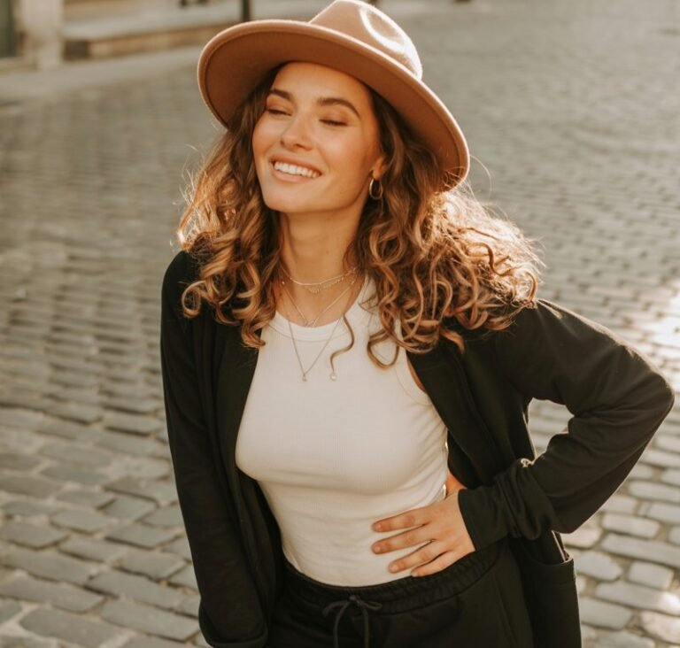 Full-body view of a white-skinned model in black joggers, white fitted top, and a tan wide-brim hat. Standing on a cobblestone street in late afternoon sunlight. Hair loose, soft curls, subtle natural makeup, layered necklaces. Pose tilting hat slightly, playful smile, one hand on hip. Emphasize authenticity, hair movement, and fabric textures.