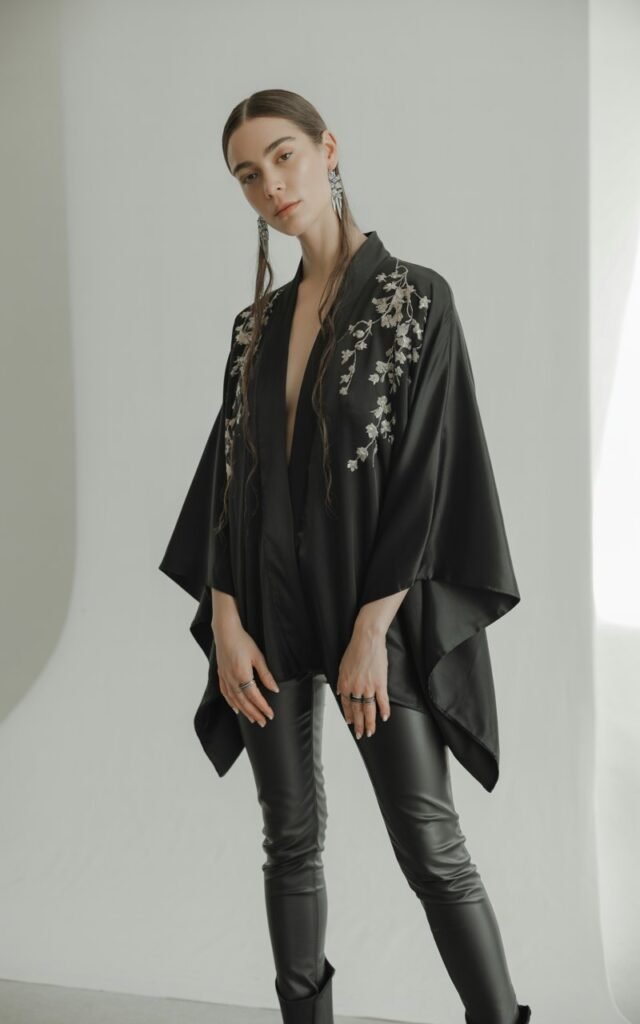 Full-body view of a white-skinned model with long dark hair, wearing a flowing black kimono with embroidered details over black leather skinny pants and black ankle boots. Accessories statement earrings, subtle rings. Pose standing in a studio with minimalistic background, arms slightly open to show kimono. Lighting soft studio lighting highlighting textures. Expression serene, head slightly tilted, natural editorial look.