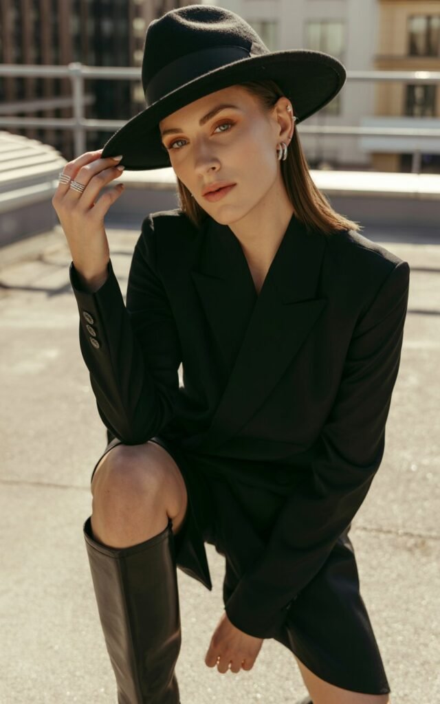 Full-body view of a white-skinned model with straight brunette hair, wearing a fitted black outfit, wide-brim black fedora, and knee-high statement boots. Accessories silver rings, minimalist earrings. Pose standing on an urban rooftop, hands lightly on hat brim. Lighting soft golden hour, subtle shadows highlighting boots. Expression confident and commanding, natural skin texture, subtle editorial makeup.