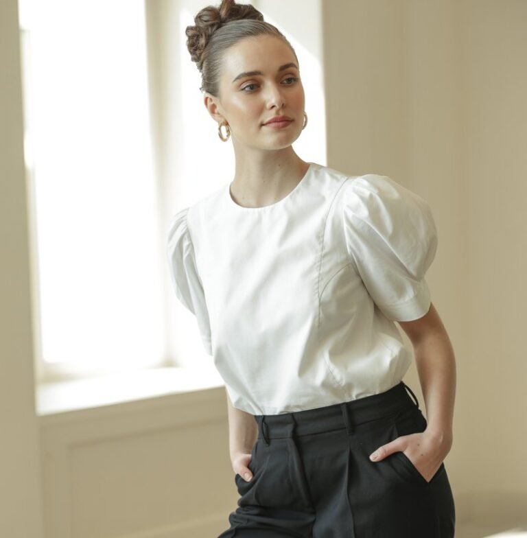 White-skinned model with elegant updo poses in an airy indoor space. She wears a puff-sleeve white blouse tucked into cropped black trousers, paired with small gold hoops and heeled mules. Soft window light illuminates her outfit. Her expression is composed yet friendly, hands gently in pockets.