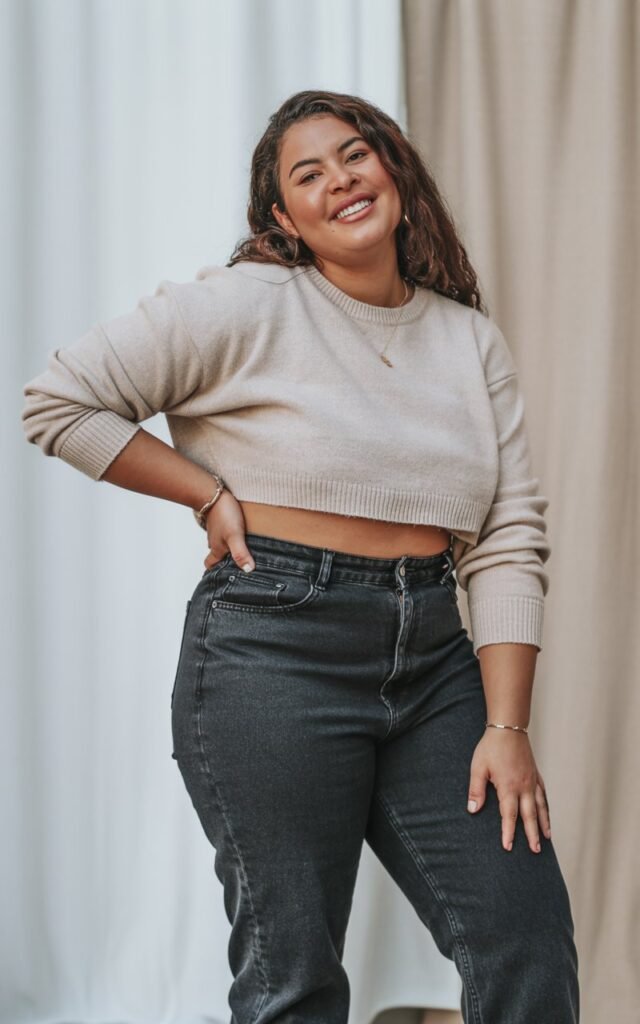 A natural-looking curvy young woman wearing high-waisted jeans with a cropped sweater. Theme curvy outfits.