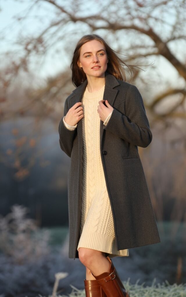 A natural-looking young woman wearing a long wool coat paired with a neutral knit dress and classic boots. Theme cold weather outfits