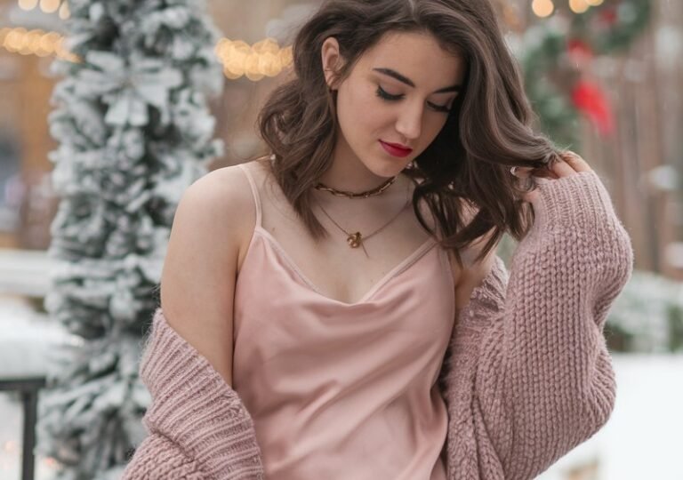 A natural-looking young woman wearing a soft pink satin slip dress styled with a chunky knit cardigan and ankle boots. Theme pink christmas outfit