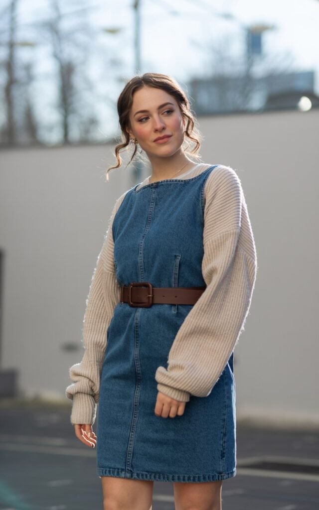 A natural-looking young woman wearing a jean skirt layered under a belted sweater dress for a winter look. Theme jean skirt outfits winter