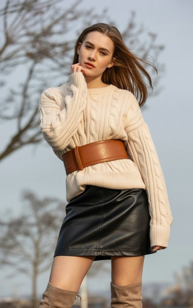 A natural-looking young woman wearing a leather skirt styled with a sweater cinched at the waist using a belt. Theme leather skirt outfit winter