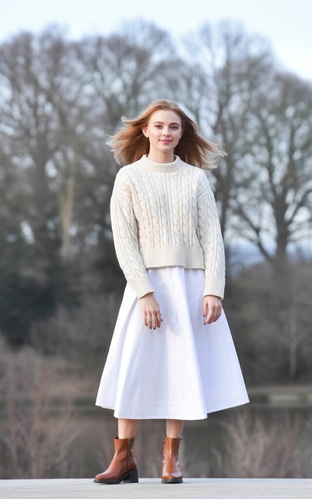 A natural-looking young woman wearing a lightweight sweater tucked into a high-waisted white skirt with ankle boots. Theme white skirt outfit winter