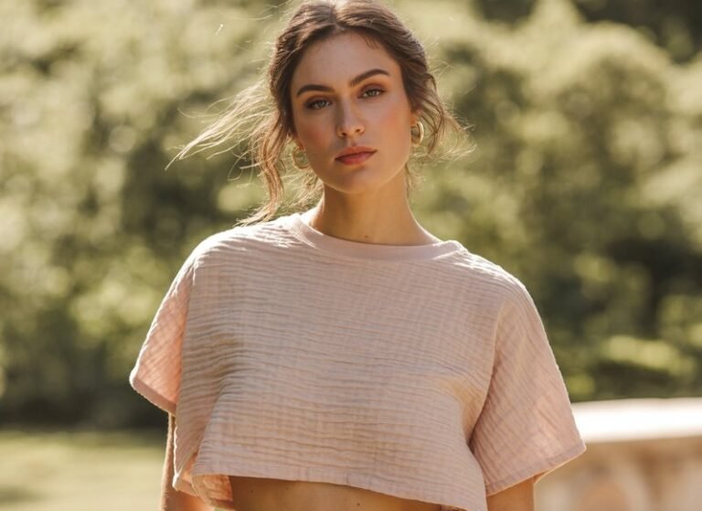A fashion photograph of a young woman with natural makeup and flowing hair, wearing a soft pastel crop top in a relaxed fit that's gently tucked into high-waisted paper-bag shorts. The shorts feature a gathered waistband with a subtle tie detail, paired with strappy leather sandals that complement the casual-chic aesthetic. She stands in a sun-drenched outdoor setting with soft, diffused natural lighting that creates a warm, effortless mood. The background features neutral tones and gentle bokeh, emphasizing the relaxed summer styling and natural beauty of the crop top ensemble.