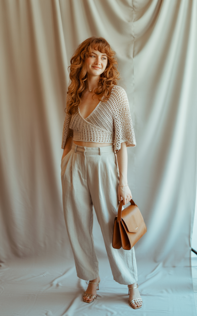 A natural-looking young beautiful woman wearing a crochet wrap top paired with high-rise linen trousers, heeled sandals, a structured handbag, and softly styled curls.