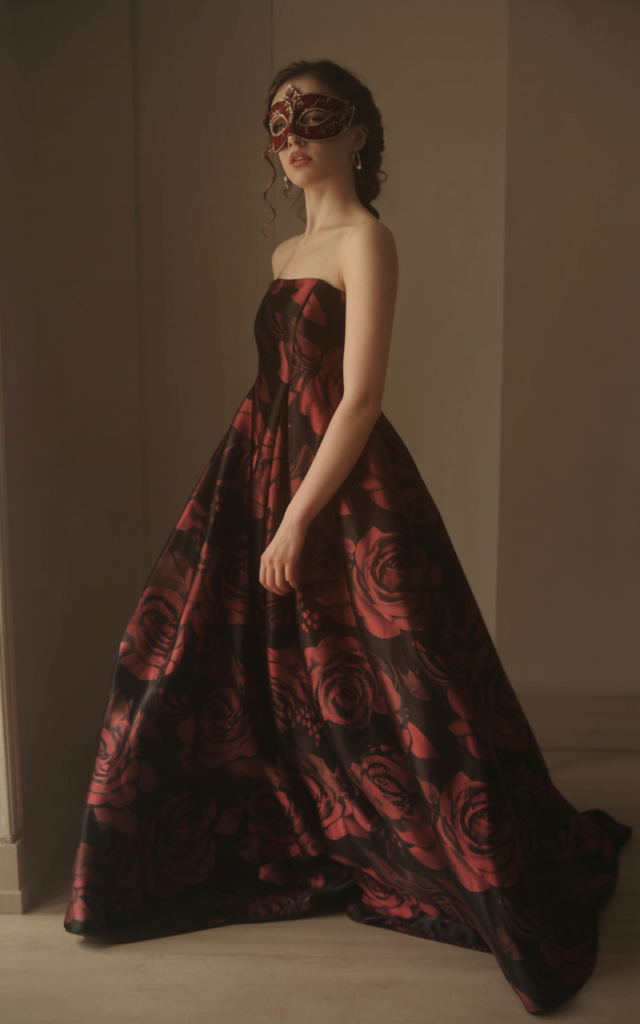 A natural-looking young beautiful woman wearing a dark floral floor-length gown with black fabric and deep red blooms, a coordinating masquerade mask, minimal accessories, and soft romantic curls.