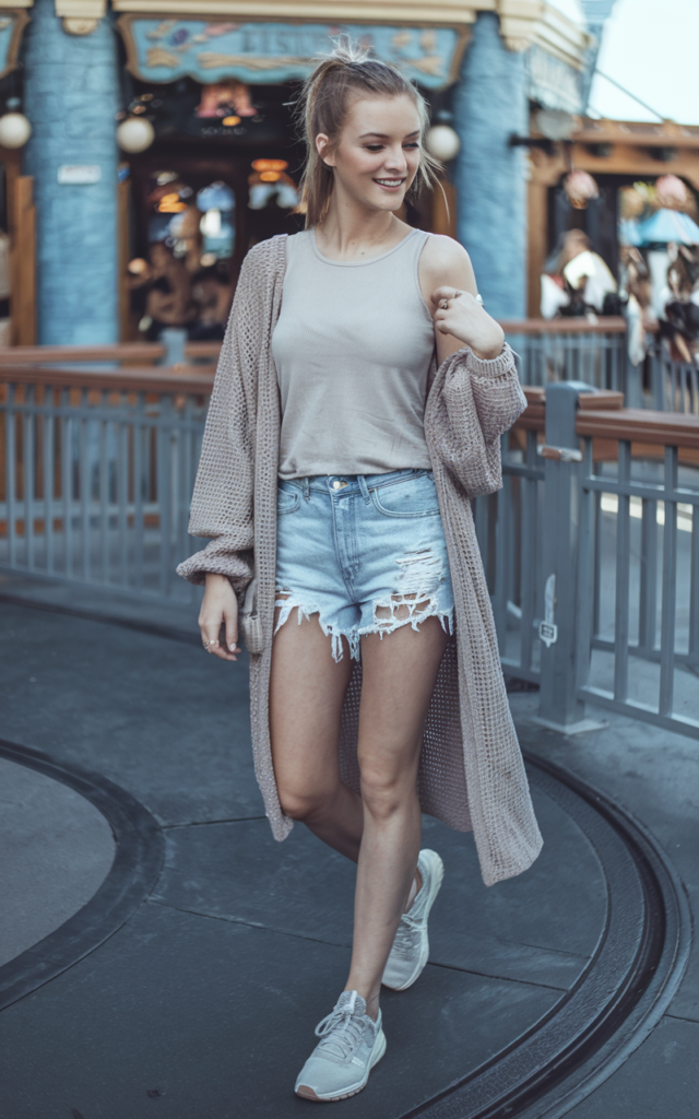 A natural-looking young beautiful woman wearing distressed denim shorts with a basic tee layered under a lightweight cardigan, white sneakers, natural makeup, and a casual ponytail at a summer Disney park.