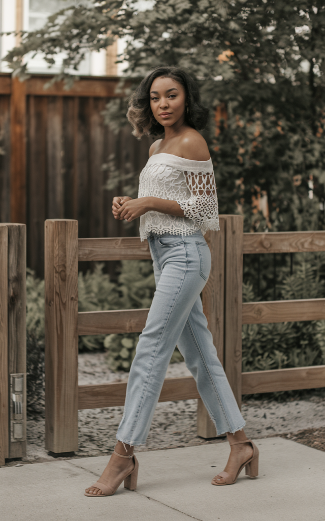 A natural-looking young beautiful woman wearing an off-shoulder crochet top with medium-wash straight-leg jeans, heeled sandals, and softly curled hair.