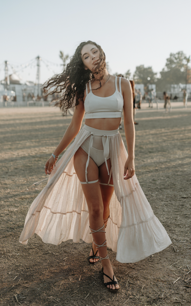 A natural-looking young beautiful woman wearing a sheer flowing maxi skirt layered over high-waisted briefs and a structured crop top, flat gladiator sandals, loose textured hair, and radiant festival makeup.