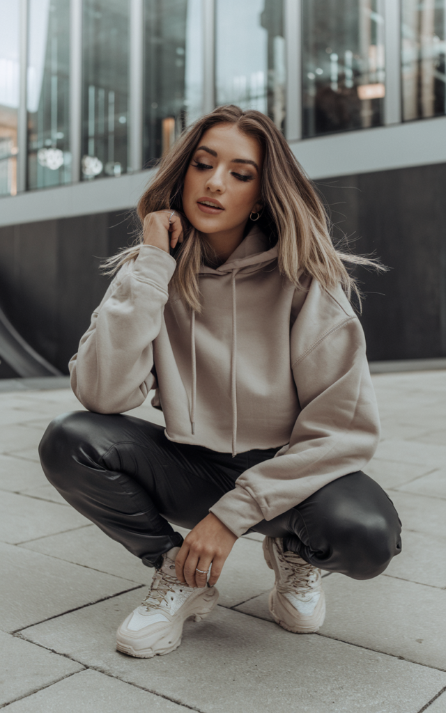 A natural-looking young beautiful woman wearing a cropped hoodie paired with slim faux leather pants, chunky sneakers, loose hair, and subtle glam makeup, styled for a Lauren Spencer Smith concert.