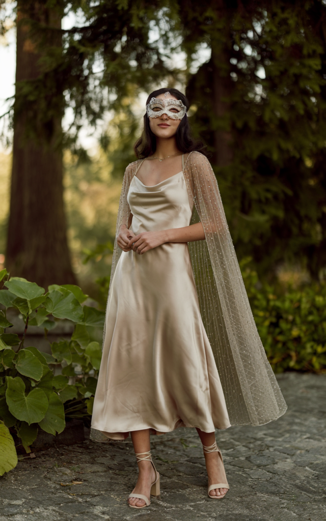 A natural-looking young beautiful woman wearing a champagne satin slip dress layered with a sheer beaded cape, a delicate masquerade mask, strappy heels, and minimal jewelry.