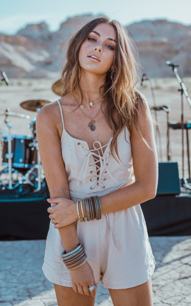 A natural-looking young beautiful woman wearing a lace-up cotton romper, western boots, stacked bangles, soft wavy hair, and sun-kissed bronzed makeup in a desert concert backdrop.