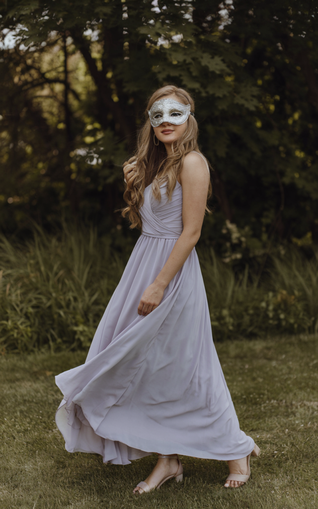 A natural-looking young beautiful woman wearing a flowing pastel chiffon gown in soft lavender, a light silver masquerade mask, loose romantic waves, minimal natural makeup, and delicate heels.