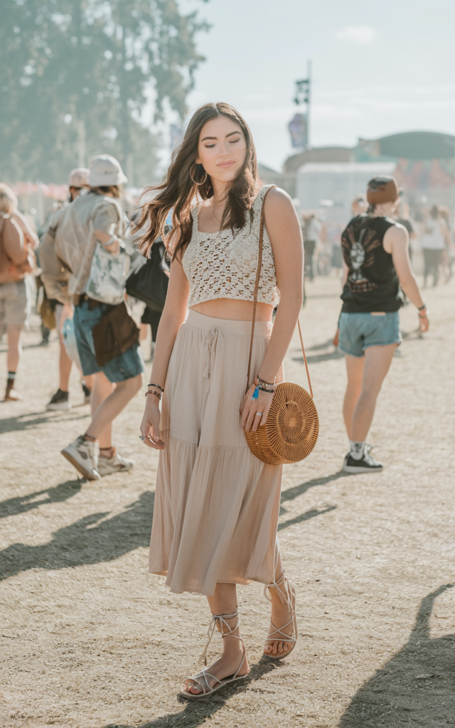 A natural-looking young beautiful woman wearing a neutral-toned crochet crop top with a matching midi skirt co-ord set, lace-up sandals, a woven crossbody bag, mixed metal jewelry, and soft glowing makeup at a sunny festival.