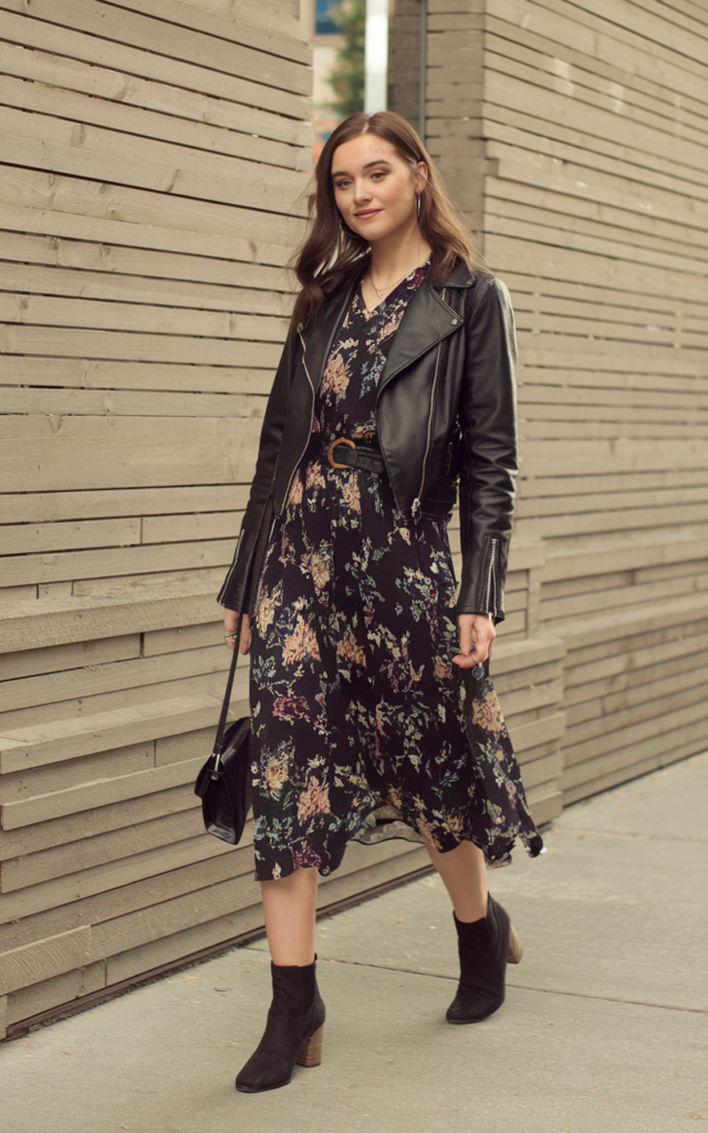 A natural-looking young beautiful woman wearing a flowy floral midi dress layered with a cropped black leather jacket, slim waist belt, heeled ankle boots, and a crossbody bag, with soft waves and subtle makeup, styled for a Lauren Spencer Smith concert.