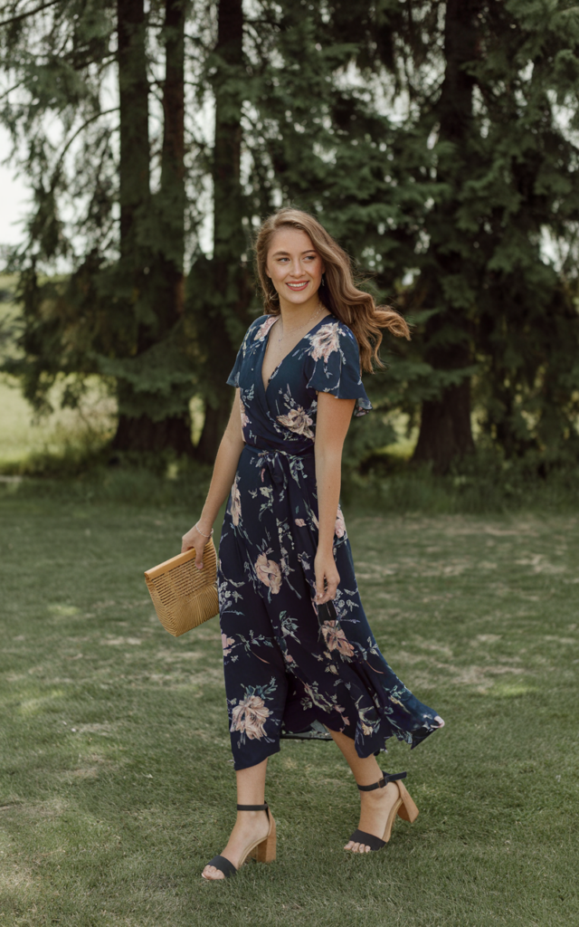 A natural-looking young beautiful woman wearing a navy floral wrap dress with soft flowing fabric, block heel sandals, a woven clutch, loose wavy hair, and fresh rosy makeup.