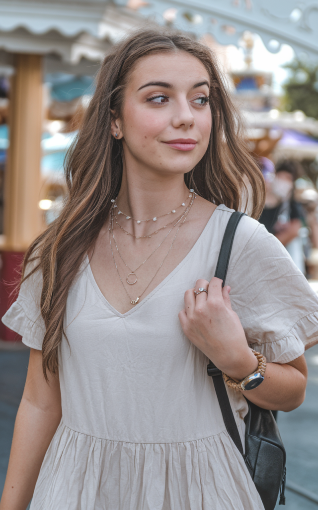 A natural-looking young beautiful woman wearing a lightweight flowy cotton sundress that moves in the breeze, cushioned white sneakers, delicate layered necklaces, and a mini backpack, with soft natural makeup and loose summer waves at a Disney park.